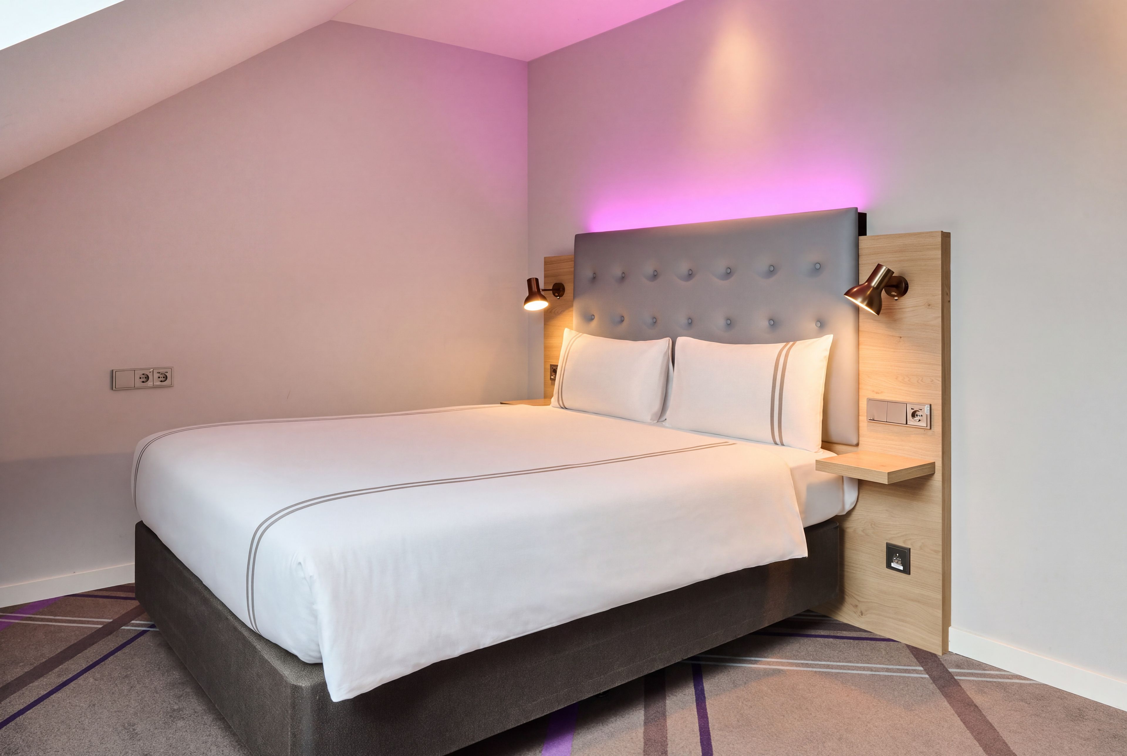 A modern hotel room with a large bed, a headboard with a light fixture, and a bedside table.
