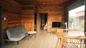 A wooden cabin interior with a sofa, a small round table, a wooden dining table with chairs, a TV, and a sliding glass door leading to an outdoor area.