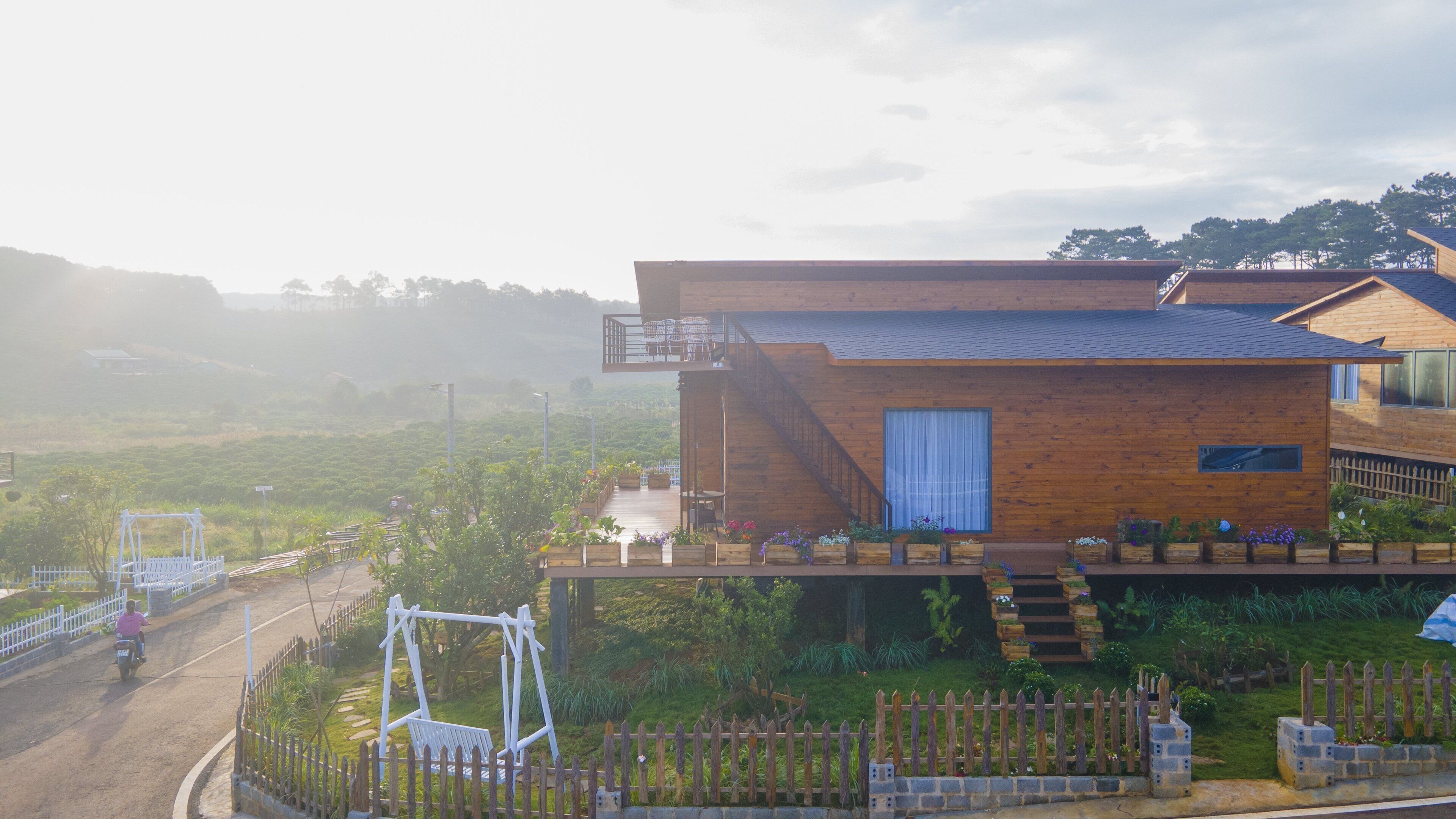 A modern wooden house with a balcony, a garden with various plants, and a swing set.