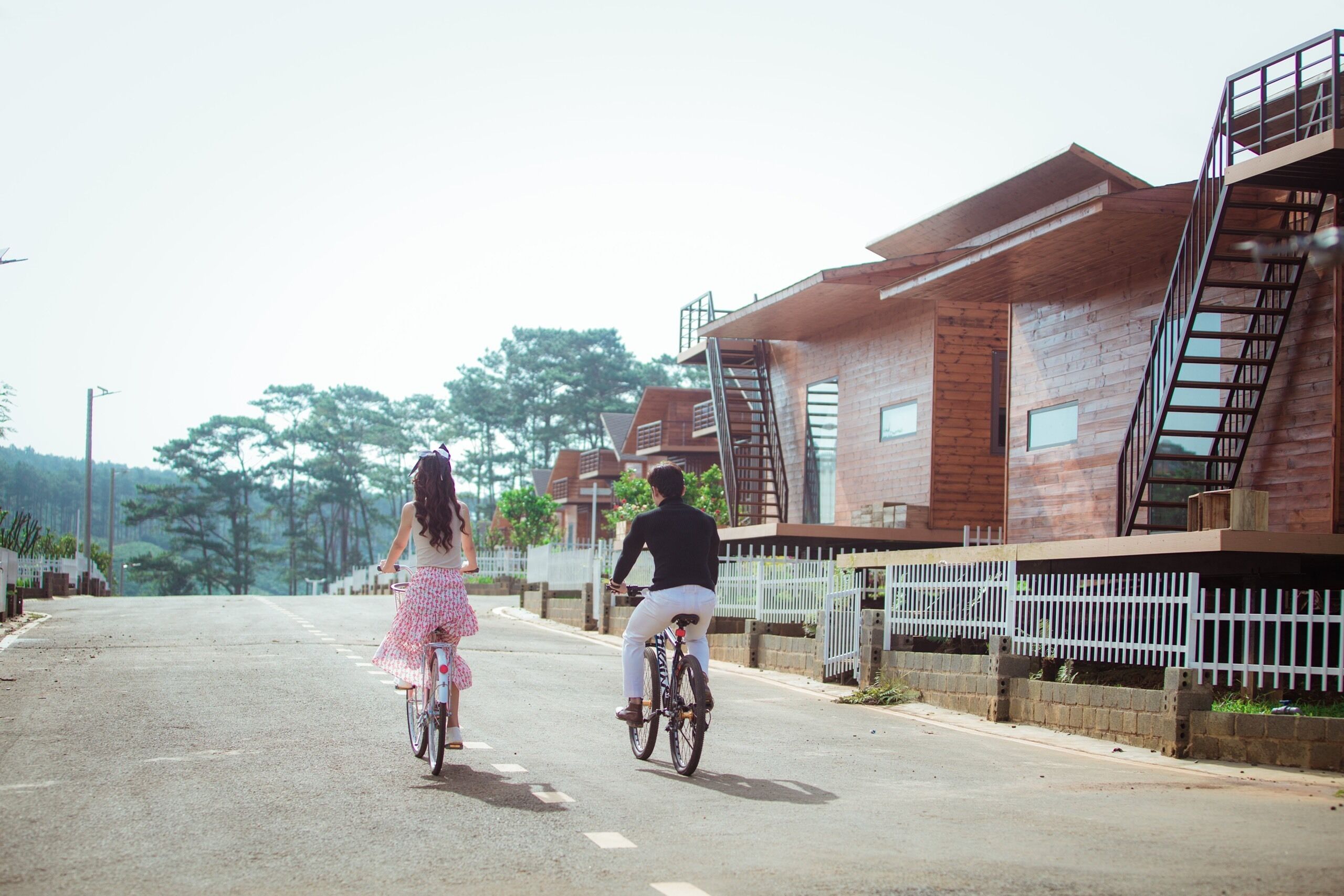 A street with two people riding bicycles, a wooden building with a staircase on the right, and a white fence along the sidewalk.