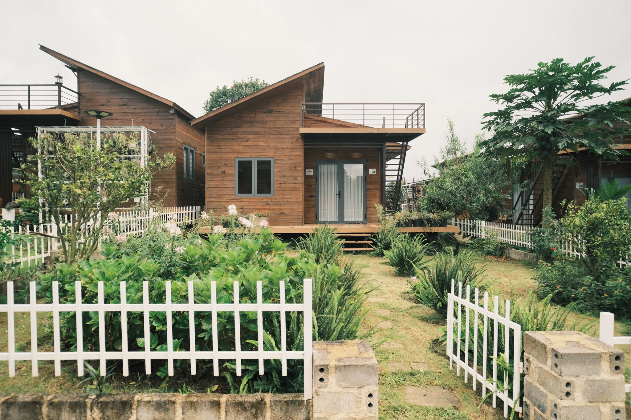 A wooden cabin with a balcony, surrounded by a white picket fence and lush greenery.