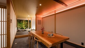 Una stanza tradizionale in stile giapponese con pavimento in tatami, un tavolo da pranzo in legno con sedie, un tavolino con un vaso e un teiera, e una porta scorrevole che dà sul giardino.