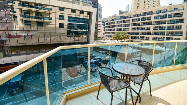 A balcony with a glass railing, two wicker chairs, and a view of a parking lot and buildings. - BRZ - Budget studio with balcony (Dubai)