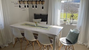 A dining area with a white table, four white chairs, a hanging light fixture, a clock, and a window with a view of trees. - Ferienhaus Hopfental (Kelheim)