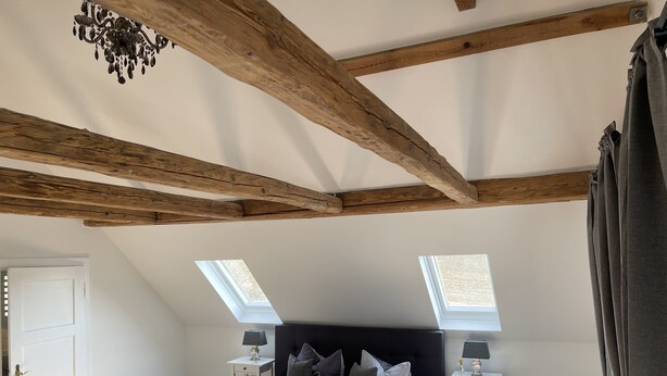 A bedroom with a sloped ceiling, wooden beams, a bed with pillows, two bedside tables with lamps, and skylights. - Ferienhaus Hopfental (Kelheim)