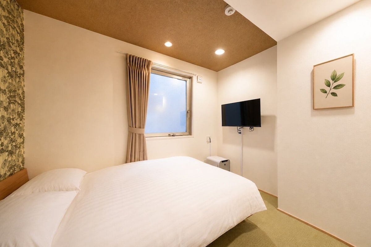Una habitación de hotel con cama, ventana con cortinas, televisor montado en la pared y un cuadro enmarcado.