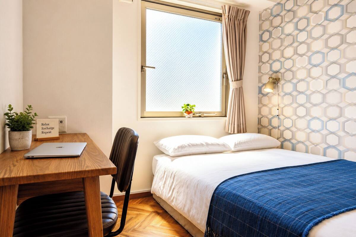 Un dormitorio con cama, escritorio con computadora portátil, una silla, ventana con cortinas, pared con diseño geométrico y una planta pequeña en el alféizar.