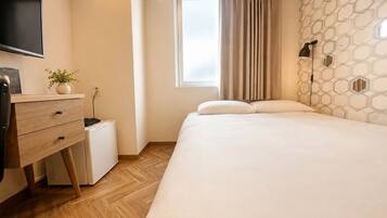 A bedroom with a bed, a desk with drawers, a small white appliance, a potted plant, a window with curtains, and a wall with a geometric pattern.