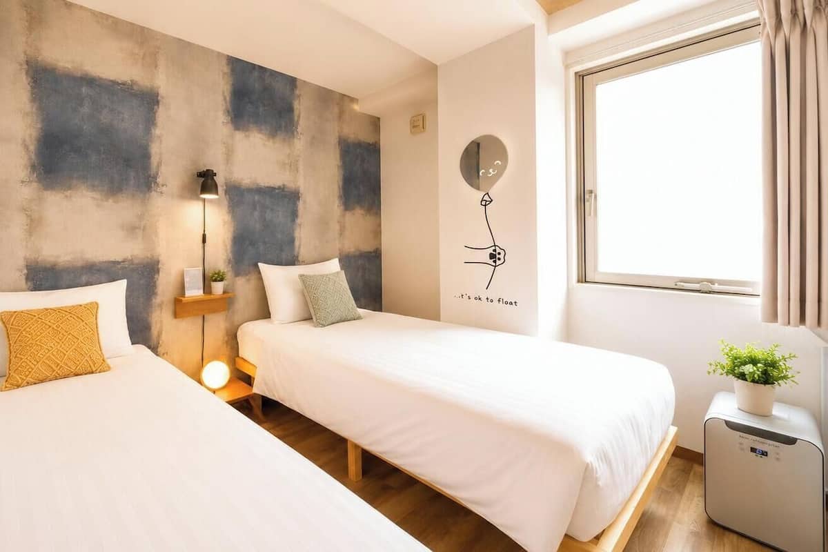 Una habitación de hotel con dos camas, una obra de arte en la pared, una lámpara de noche, una mesita con una planta y una ventana con cortinas.