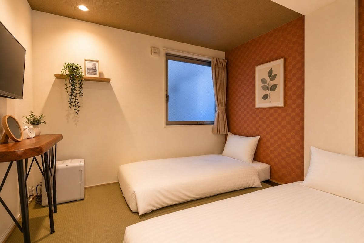 Habitación de hotel con dos camas, un escritorio pequeño con un reloj, un televisor, una ventana con cortinas y una planta en la pared.