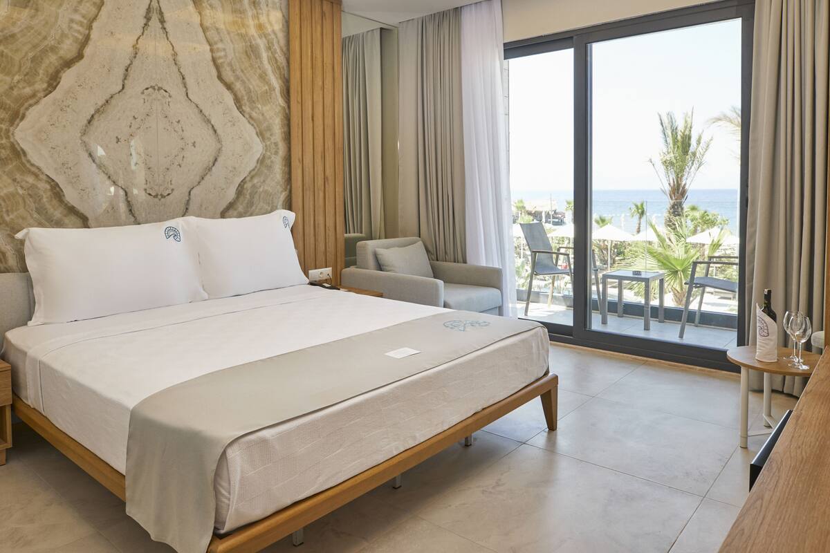 Habitación de hotel moderna con una cama grande, vista al océano, balcón con mobiliario exterior y un panel de madera decorativo.
