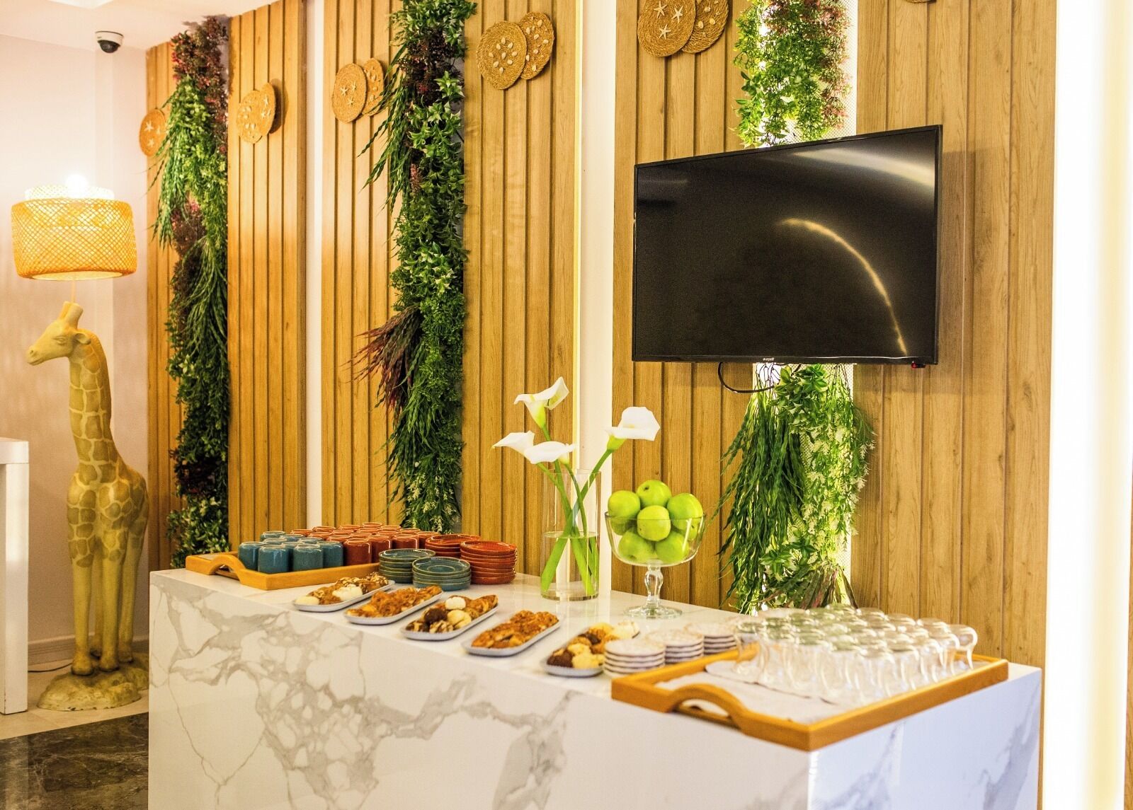 A buffet setup with various dishes, a television mounted on the wall, and decorative plants.