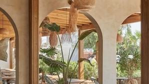 A covered outdoor seating area with wooden beams and hanging lights, surrounded by large potted plants.