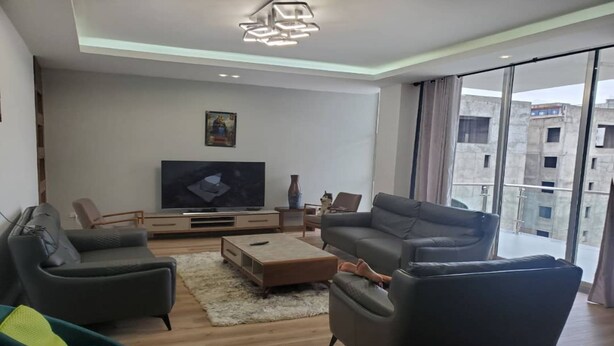 A modern living room with a flat-screen TV, grey sofas, and a large window offering a city view.