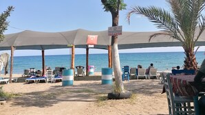 Un café situé en bord de mer, avec des tables à l’extérieur sous des parasols, un panneau indiquant un espace réservé aux fumeurs et une vue sur la mer.