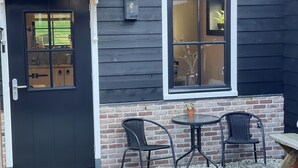 L’extérieur est en bois foncé, avec une porte noire et une fenêtre. On y trouve deux chaises en osier et une petite table.