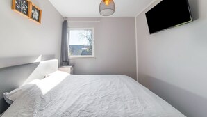 Un dormitorio con cama, televisor montado en la pared, ventana con cortinas y lámpara en el techo.