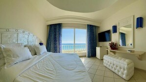 Une chambre d’hôtel avec un grand lit, un banc blanc, une télévision, un miroir et une vue sur la mer.