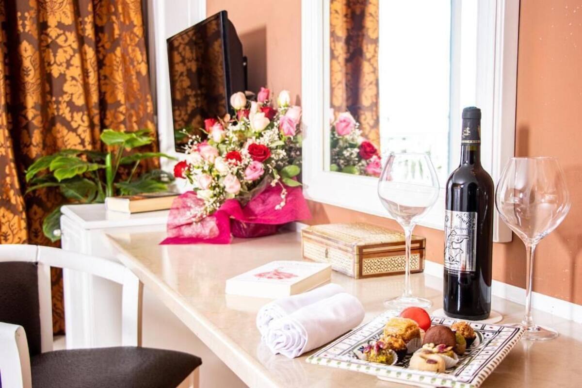 Habitació d'hotel amb zona de menjador que inclou una taula preparada amb una ampolla de vi, gots, un plat de pastissos, un ram de flors i un llibre.