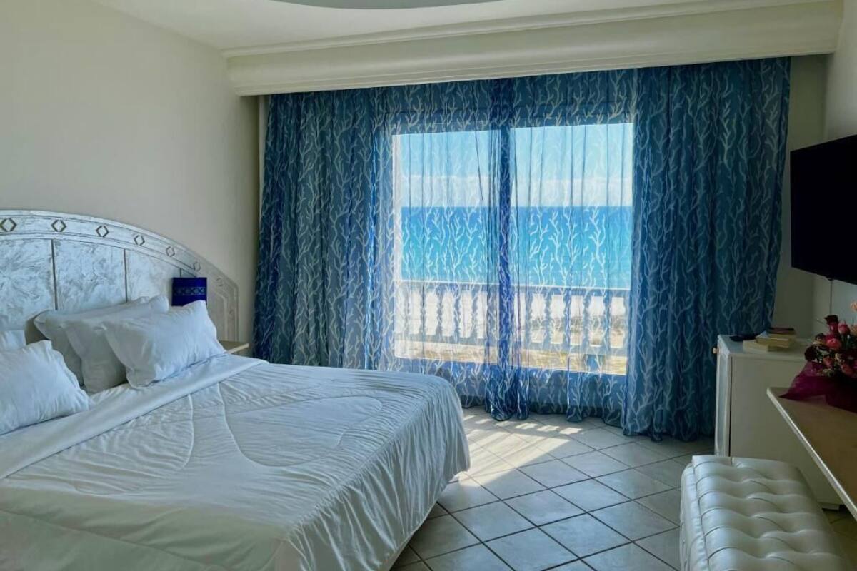 Habitació d'hotel amb llit, escriptori i vista a la platja a través de cortines transparents.