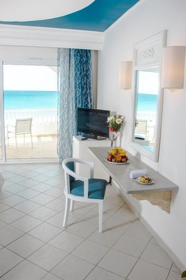 A room with a TV, a dining table with fruit, a chair, a flower vase, and a view of the beach.