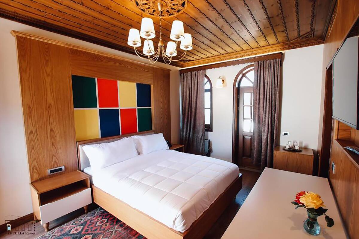 Habitación de hotel con techo de madera, cama con ropa de cama blanca, mesita de noche, una pequeña mesa con un jarrón de flores y un panel de pared colorido.