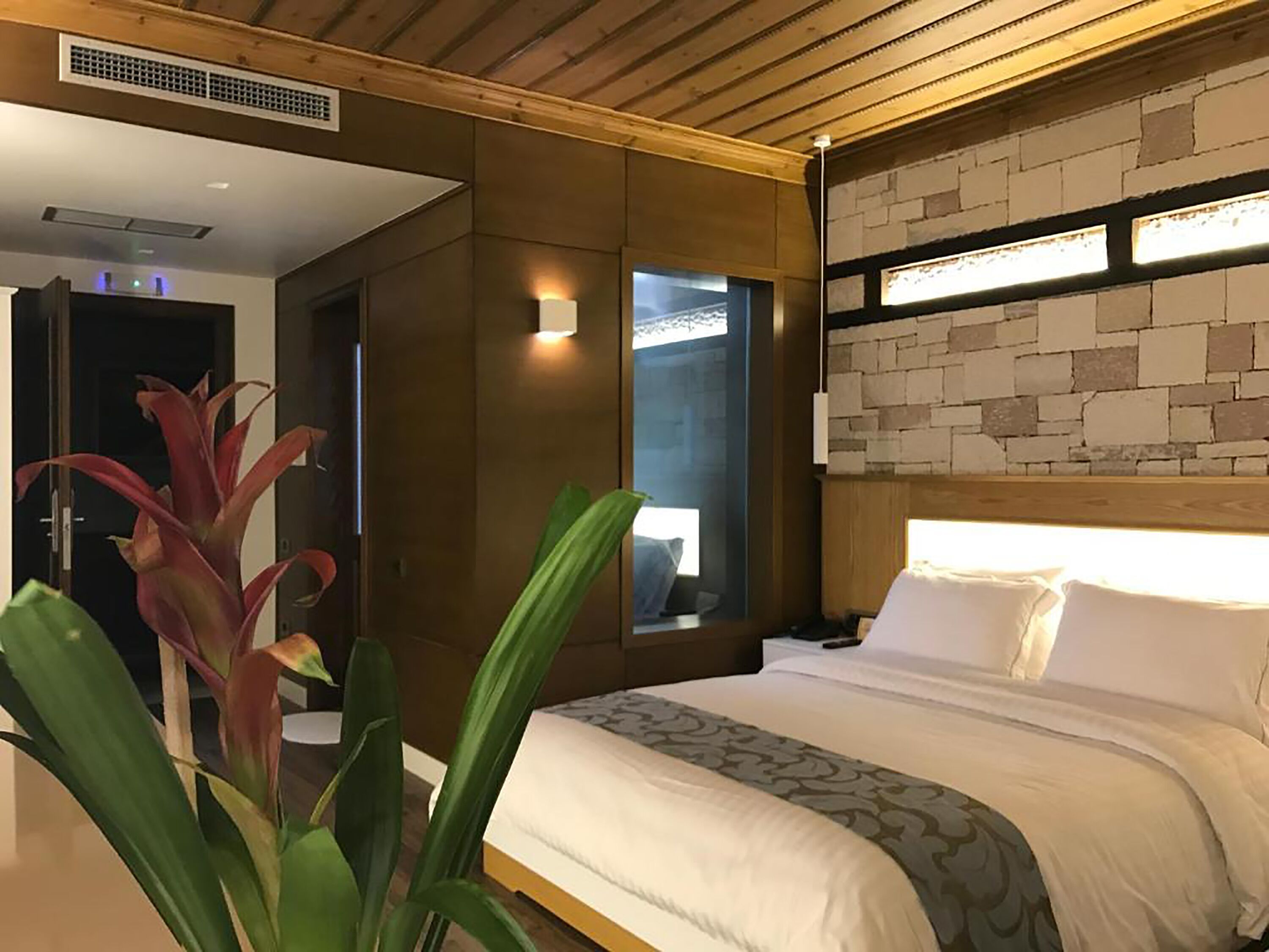 Habitación de hotel con techo de madera, pared de piedra, cama con ropa blanca y un ventanal grande.