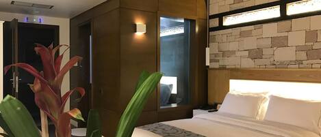 Habitación de hotel con techo de madera, pared de piedra, cama con ropa blanca y un ventanal grande.
