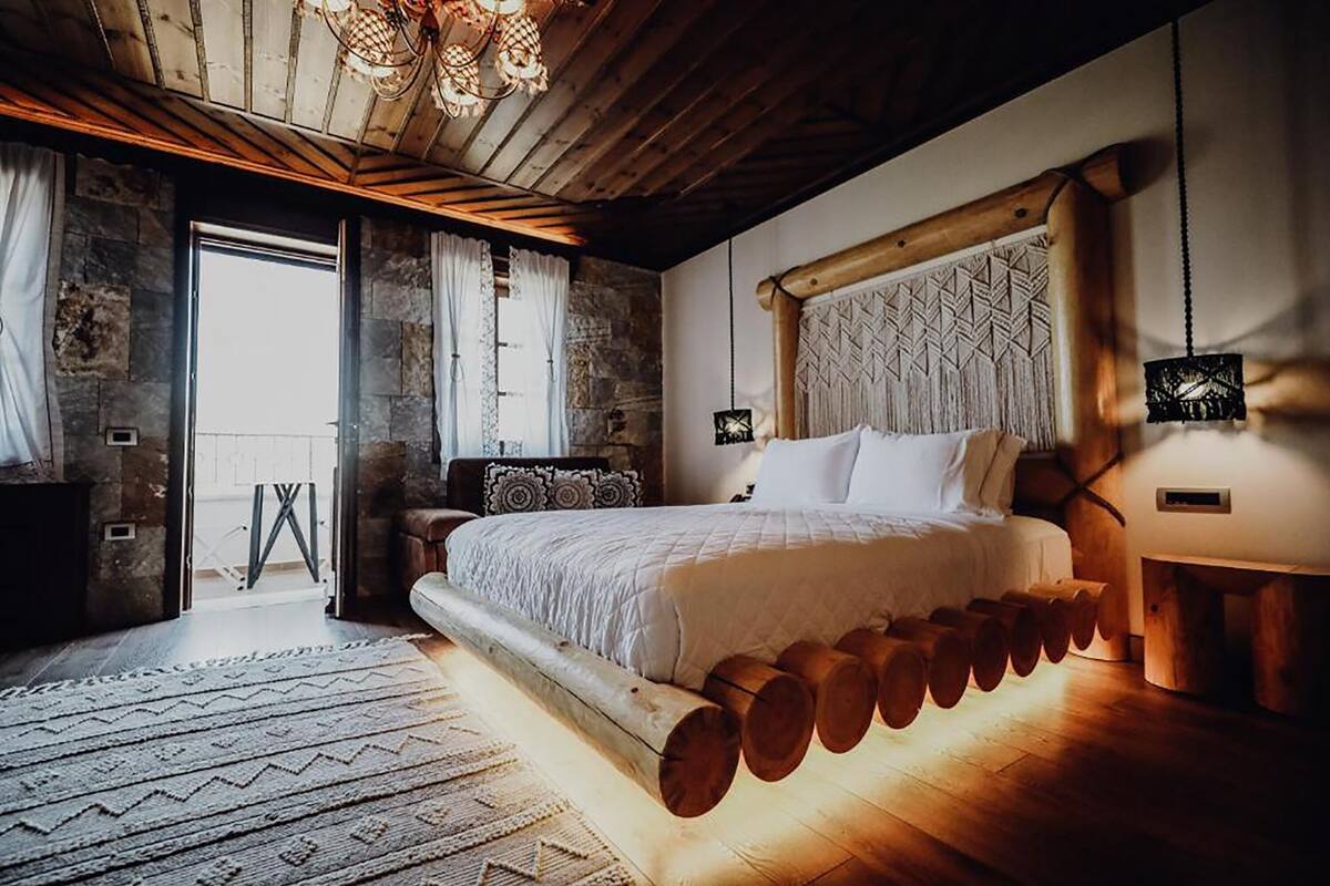 Un dormitorio con una cama grande, leños debajo de la cama, una pared de piedra y techo de madera.