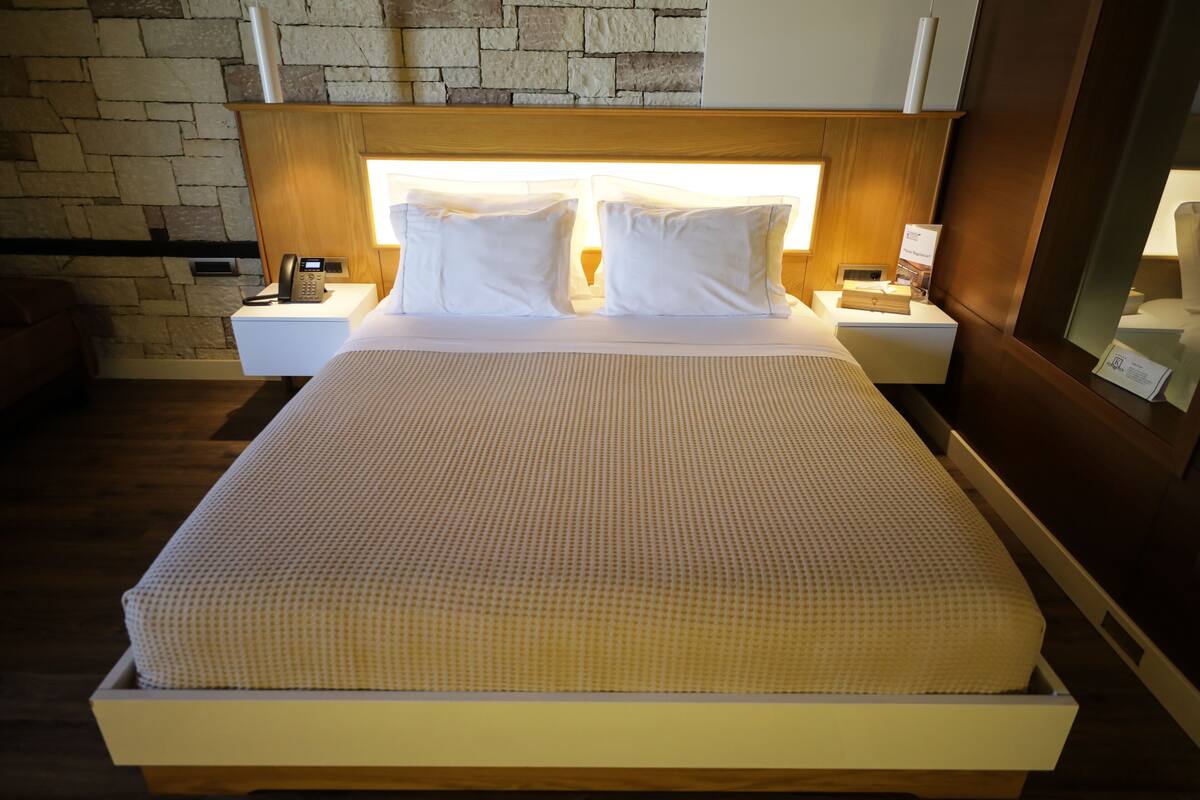 Una cama bien hecha con un cabecero, dos mesitas de noche con lámparas, y una pared de piedra al fondo.