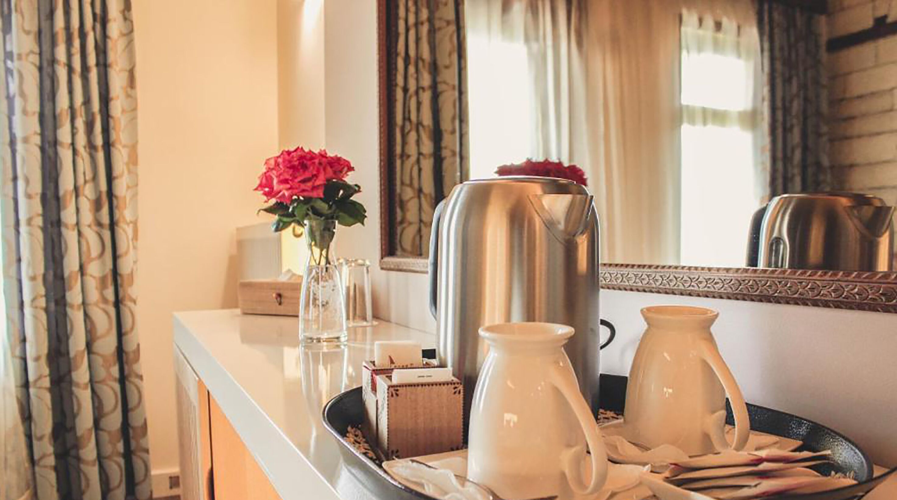 A hotel room with a white minibar, a large metallic kettle, a white pitcher, and a vase with red flowers.