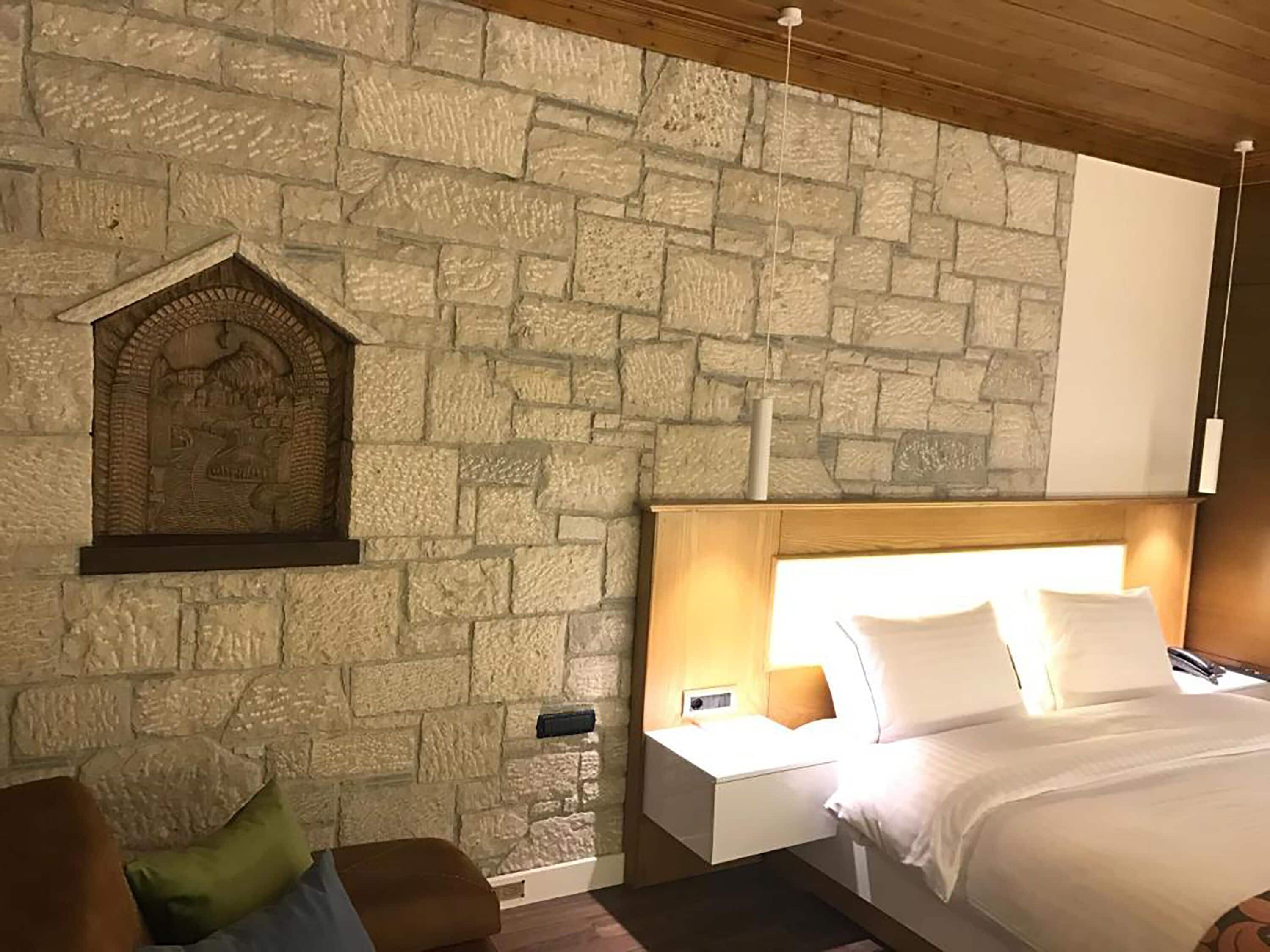 Une chambre avec un mur en pierre, un plafond en bois, un lit avec une literie blanche, une table de chevet avec une lampe et un canapé avec des coussins.