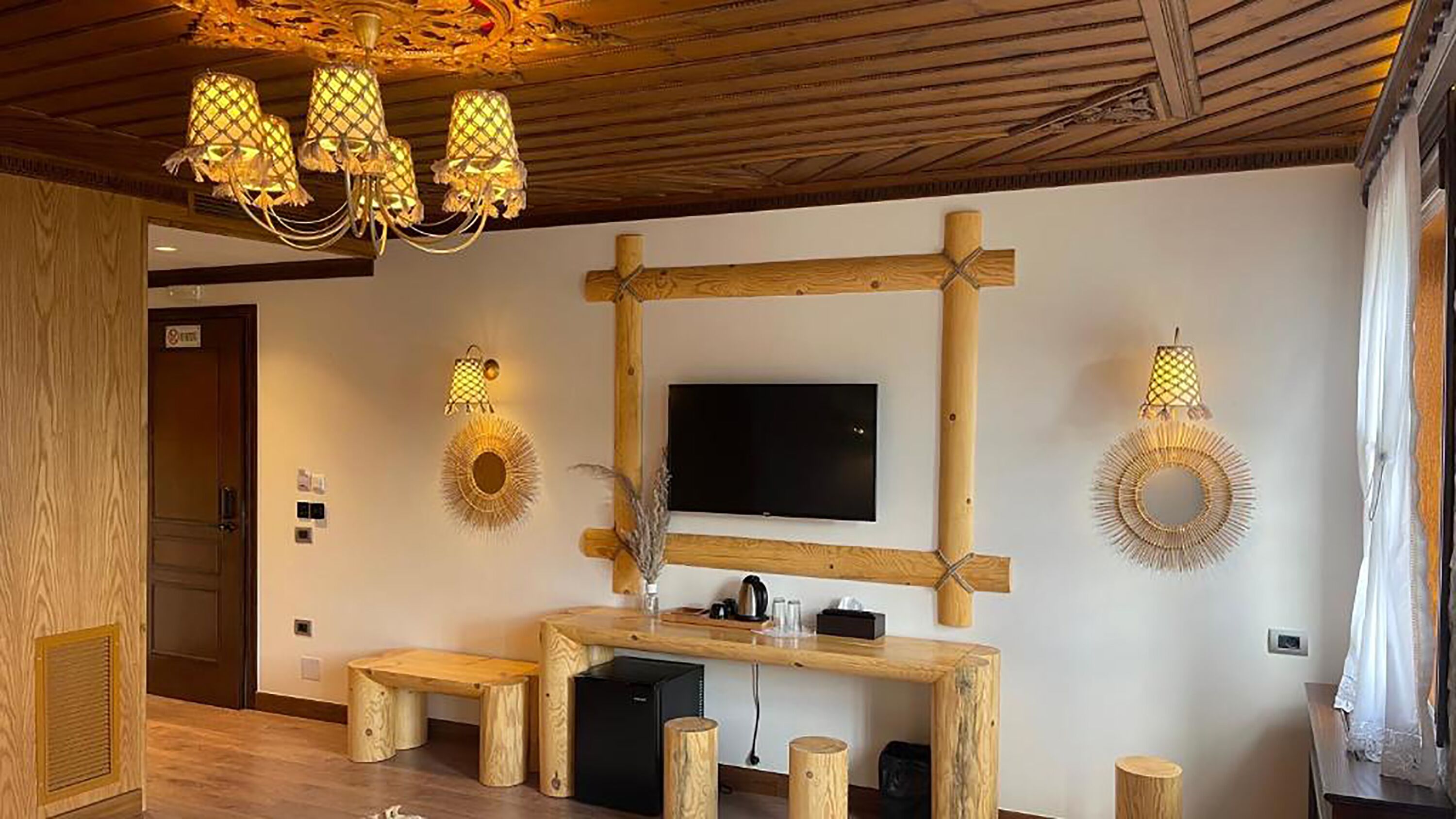 A room with a wooden ceiling, a TV mounted on a wooden frame, and a wooden bench with a small table.