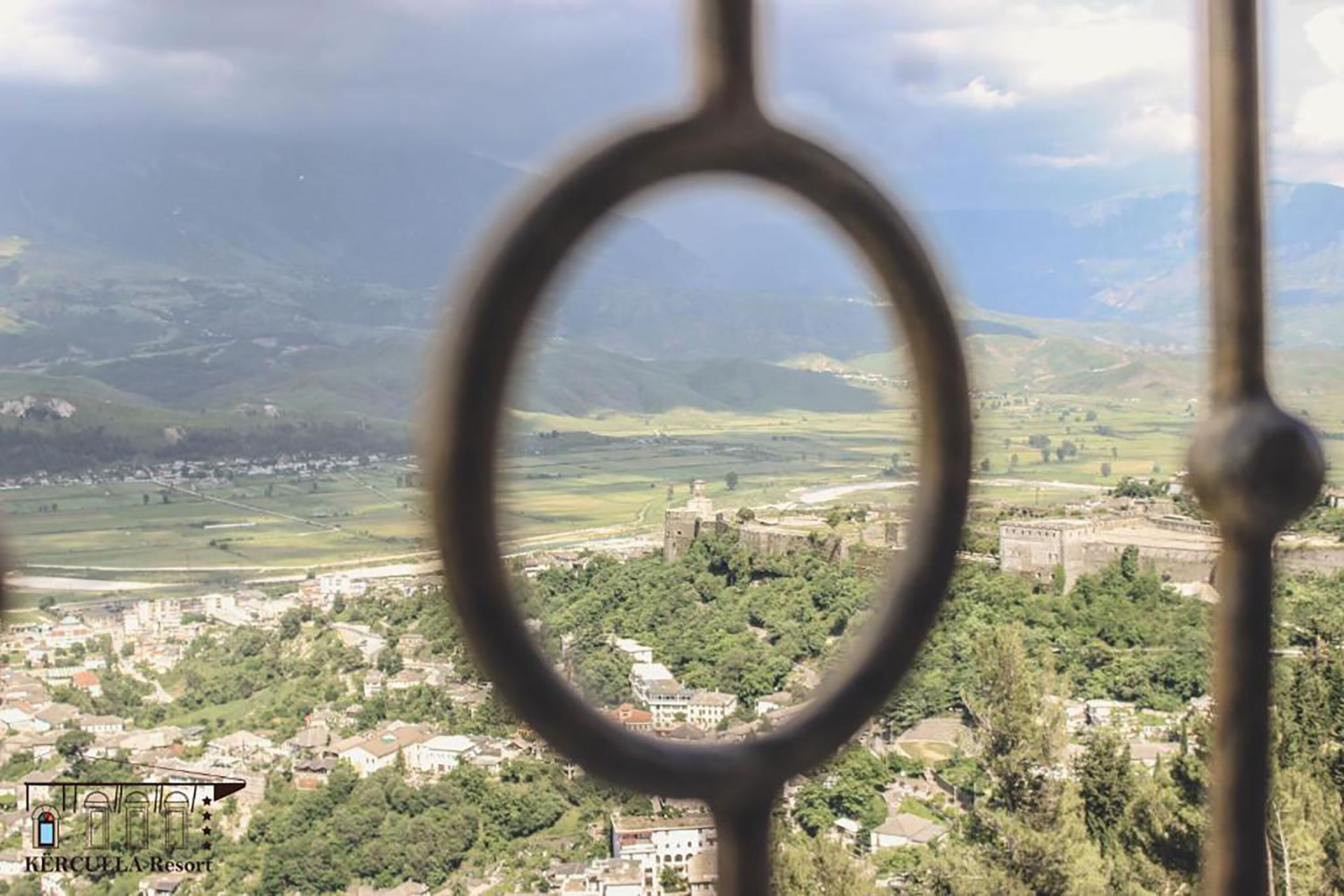 A view through a metal gate of a town with a castle, surrounded by greenery and fields.