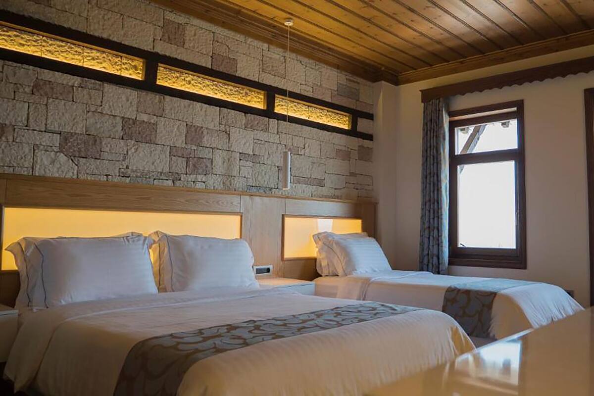 Habitación de hotel con dos camas, paredes de piedra, vigas de madera en el techo y ventanas amplias.