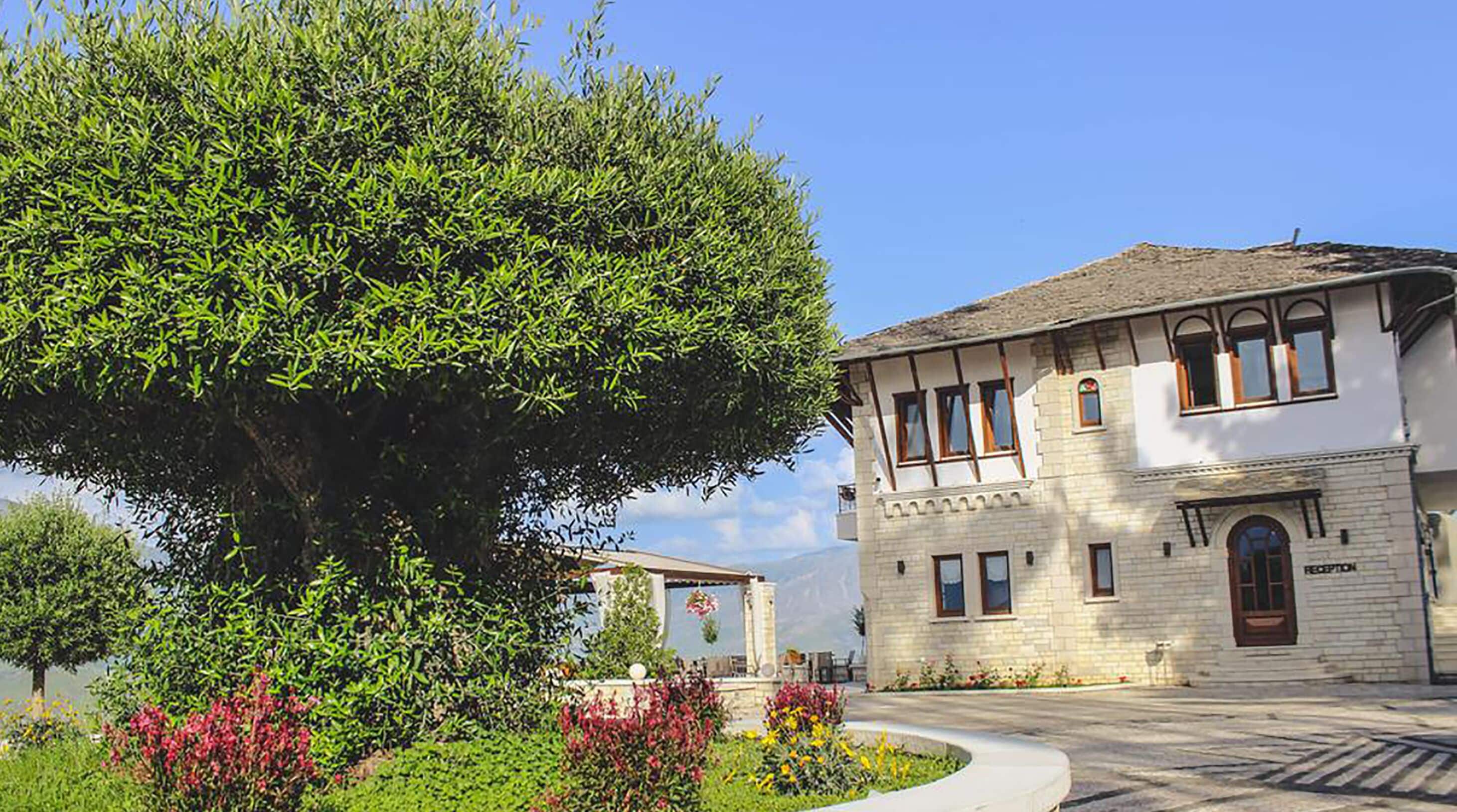 Un edificio de dos plantas con fachada de piedra, una recepción y un gran árbol en primer plano.