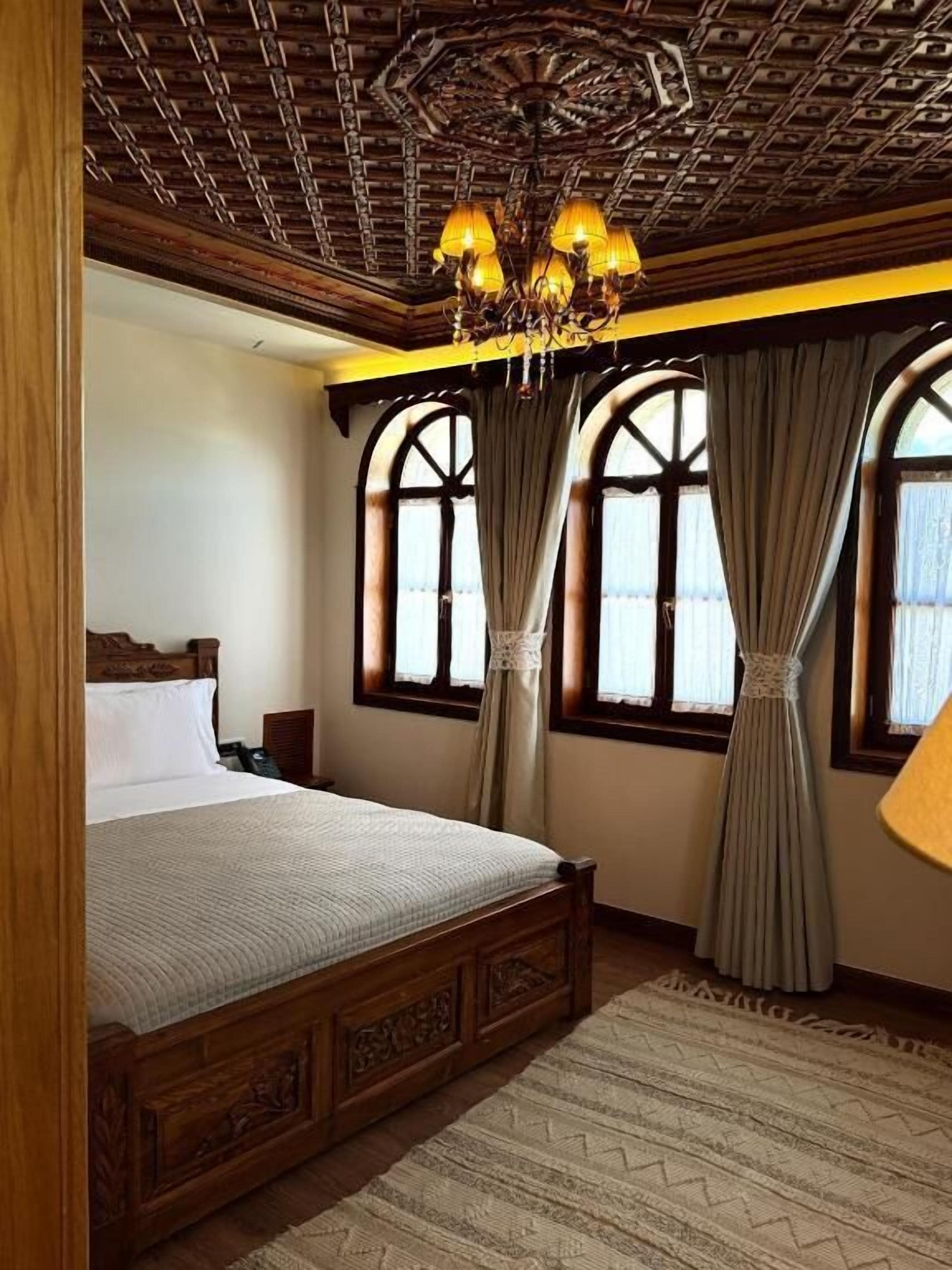 A bedroom with a large bed, wooden headboard, a chandelier, and two windows with curtains.