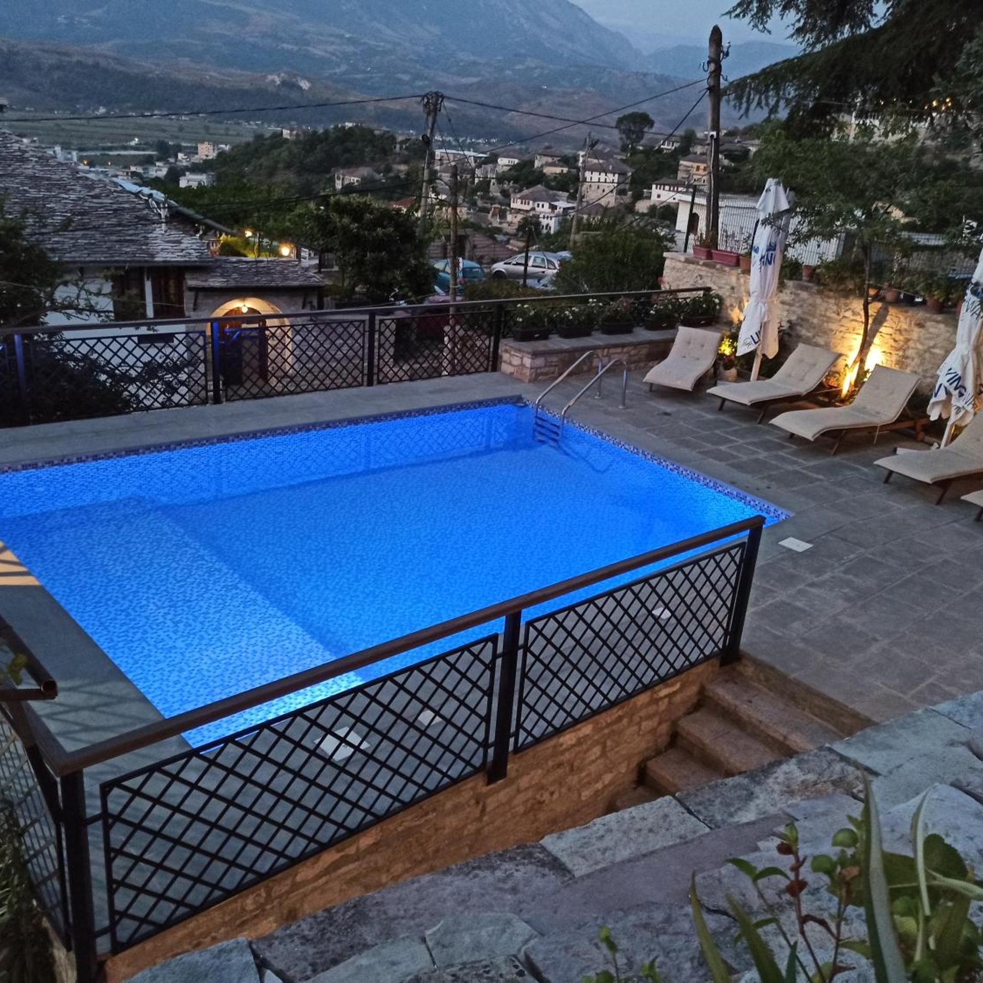 Una piscina con cerramiento metálico, sillas de descanso y vista a un pueblo y montañas al fondo.