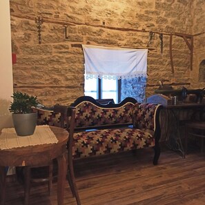A rustic interior with a stone wall, a patterned sofa, a wooden table with a potted plant, and a window with a white curtain. - Hotel Praga (Gjirokastër)