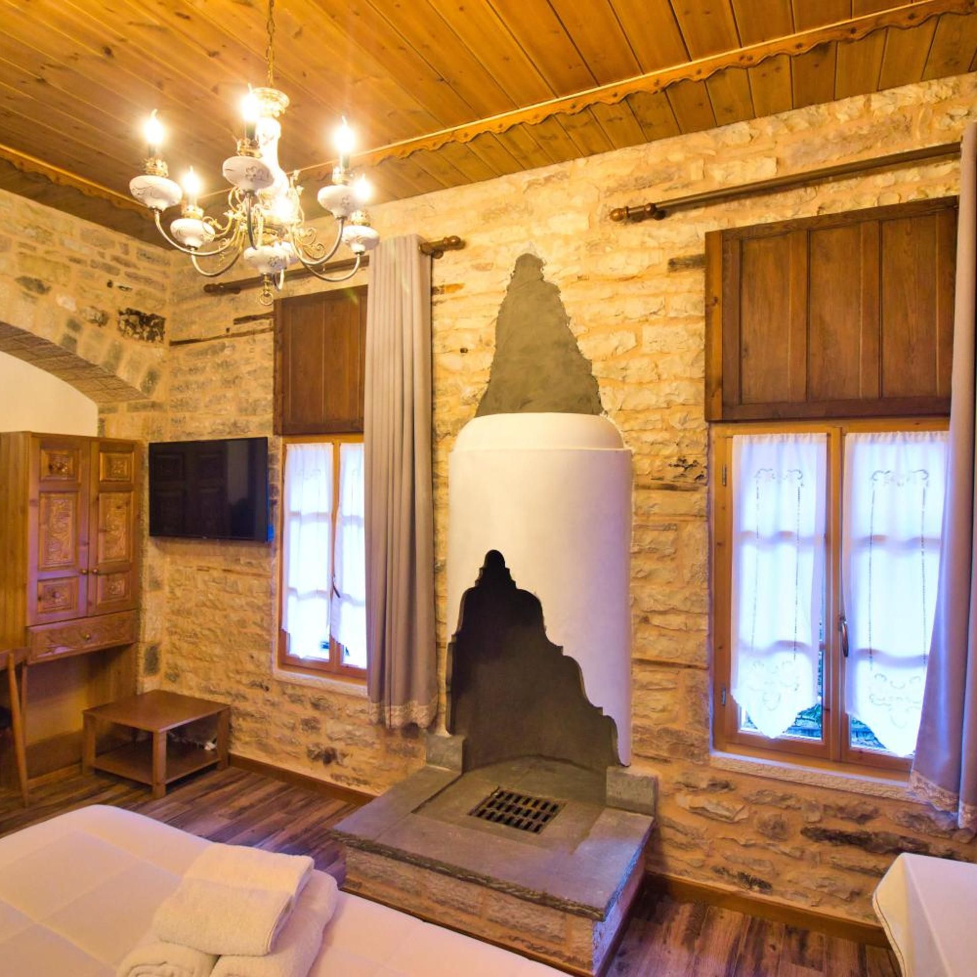A room with stone walls, a wooden ceiling, a bed with white linens, a stone fireplace, and a chandelier.