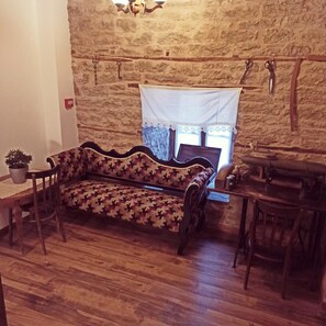 A room with a stone wall, a patterned sofa, a small table with a plant, and a window with a white curtain. - Hotel Praga (Gjirokastër)
