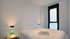 A neatly made bed with a folded towel, a bedside table with a lamp, and a window with a view of the sky. - Modern two bedroom apartment (Ronda)