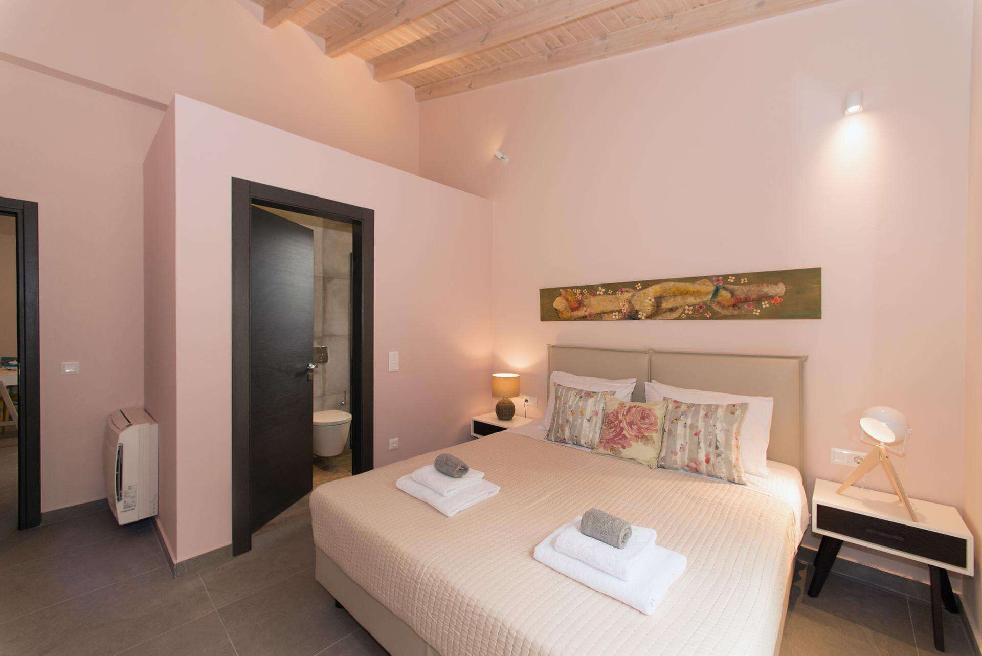 Un dormitorio con cama, mesita de noche, lámpara, un cuadro y una puerta que da al baño.