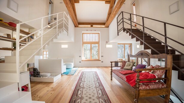 Ein geräumiges Zimmer mit einer Holztreppe, einem Bett mit gemusterten Textilien, einem weißen Sofa und einem großen Fenster.