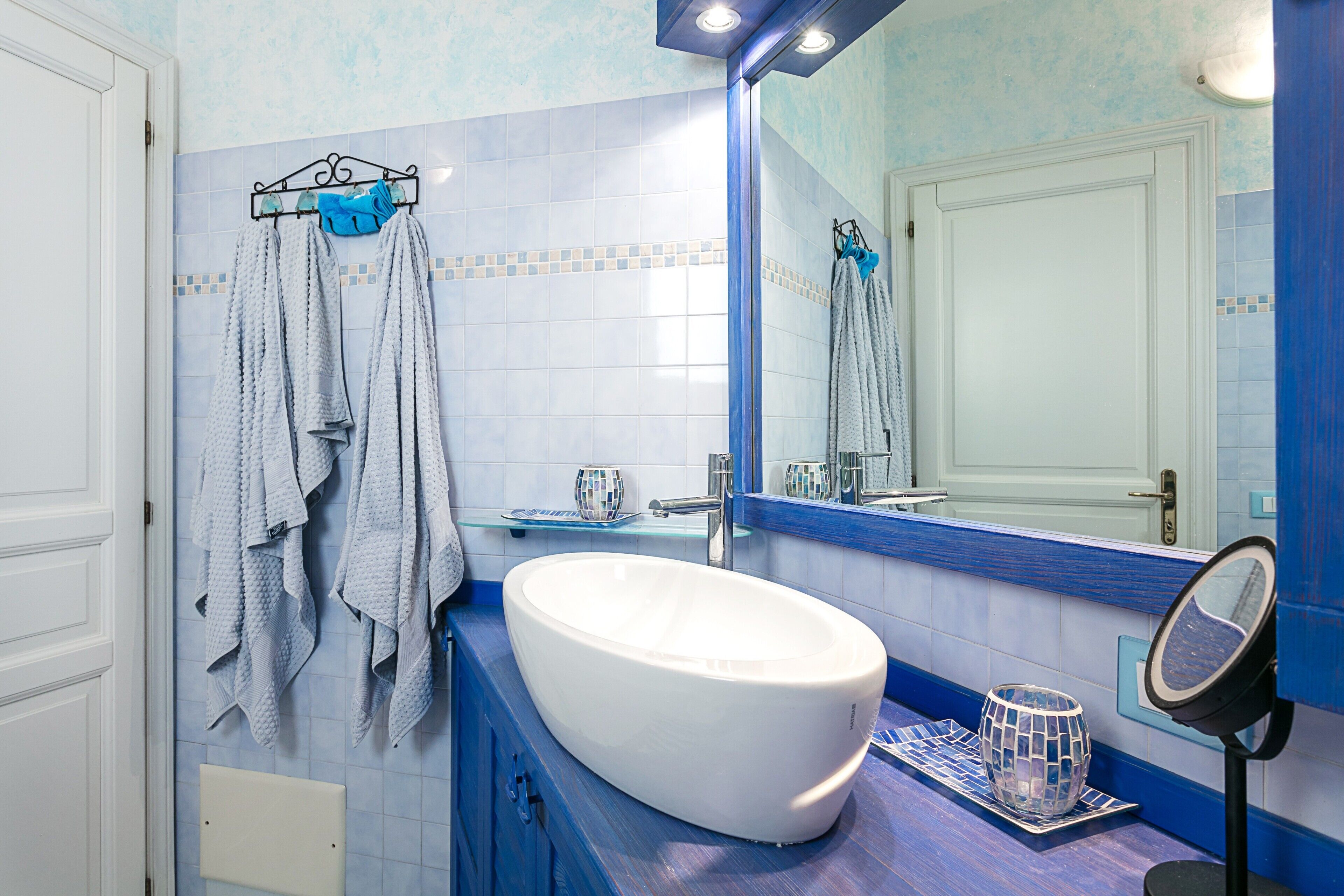 Casa de banho com uma pia branca, azulejos azuis, um espelho e toalhas penduradas num porta-toalhas.