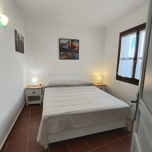 A bedroom with a bed, two bedside tables with lamps, a wall-mounted painting, and a window with curtains.