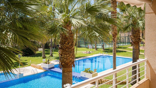 A swimming pool with palm trees, a white railing, and a view of a building in the background. - Apartment 'Alborada Golf  1A' with Terrace, Shared Pool & Wi-Fi (Alfaz del Pi)