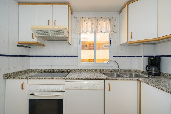 A kitchen with white cabinets, a granite countertop, a built-in oven, a dishwasher, a window with a curtain, and a coffee maker.