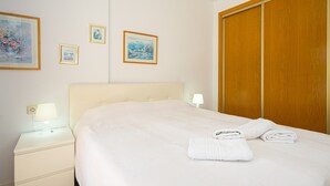 A hotel room with a bed, a nightstand with a lamp, a wooden wardrobe, and three framed pictures on the wall. - Apartment 'Alborada Golf  1A' with Terrace, Shared Pool & Wi-Fi (Alfaz del Pi)