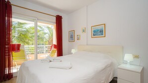 A hotel room with a bed, a nightstand with a lamp, a window with curtains, and a view of palm trees. - Apartment 'Alborada Golf  1A' with Terrace, Shared Pool & Wi-Fi (Alfaz del Pi)